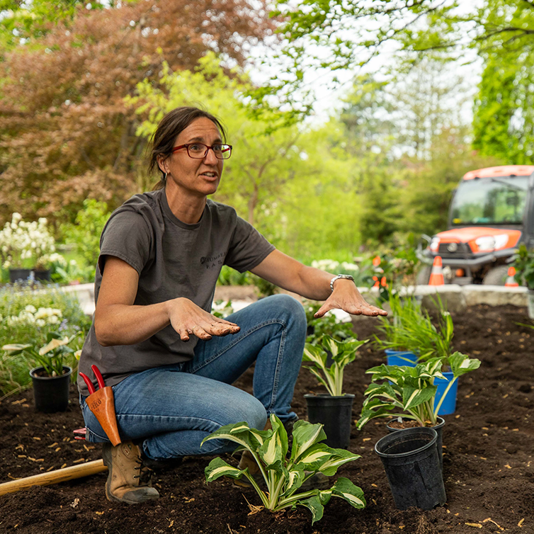 A woman kneels in a garden bed, explaining a concept to the viewer