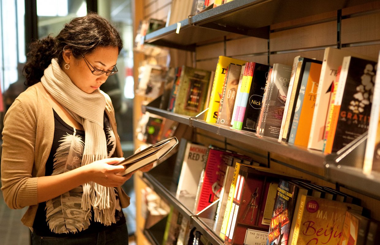 A woman wearing a white scarf stands in the library, staring at a book in her hand.