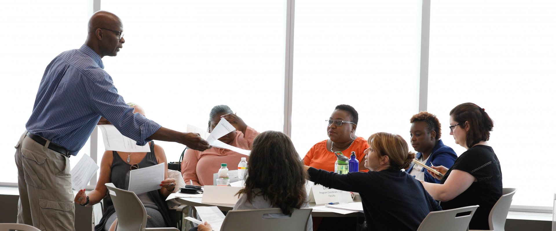 A black man hands out documents to a table of adult students