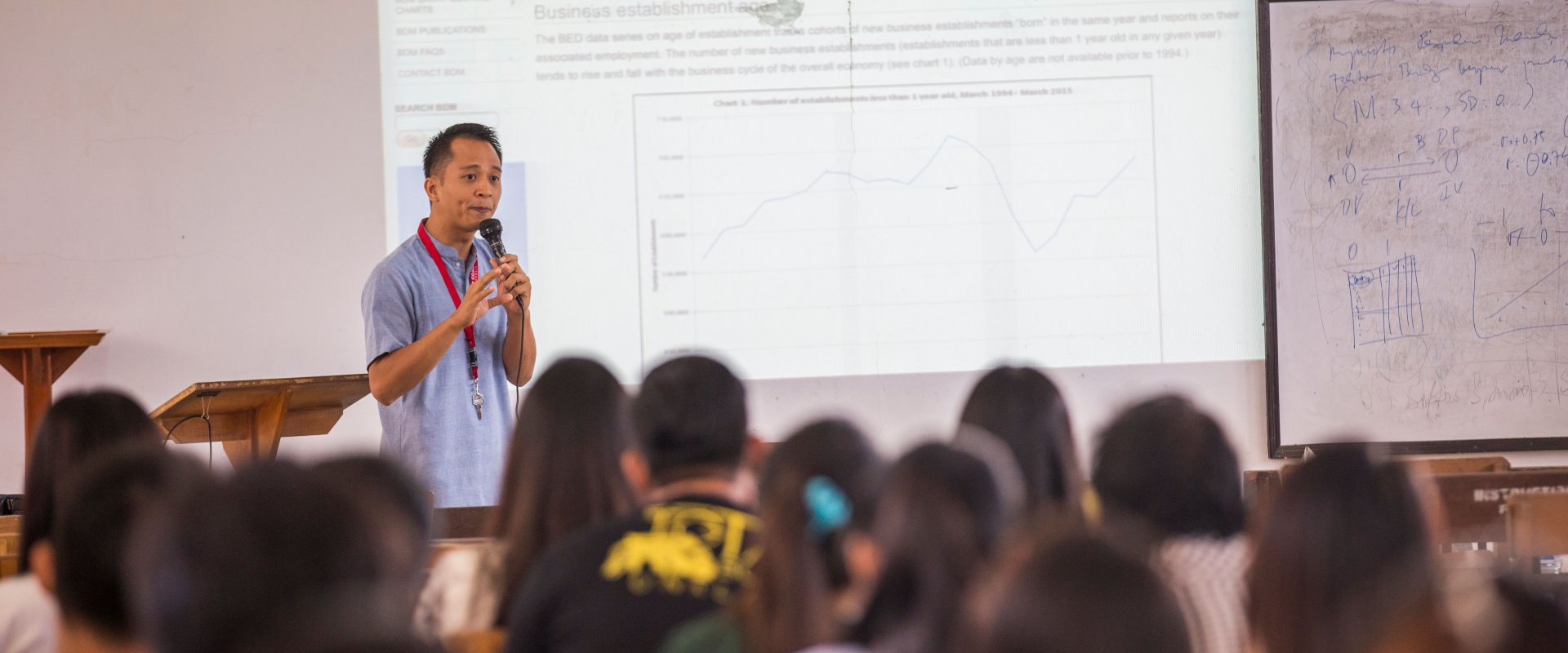 An Asian man stands at the front of a classroom full of students, present with a microphone and projection screen.