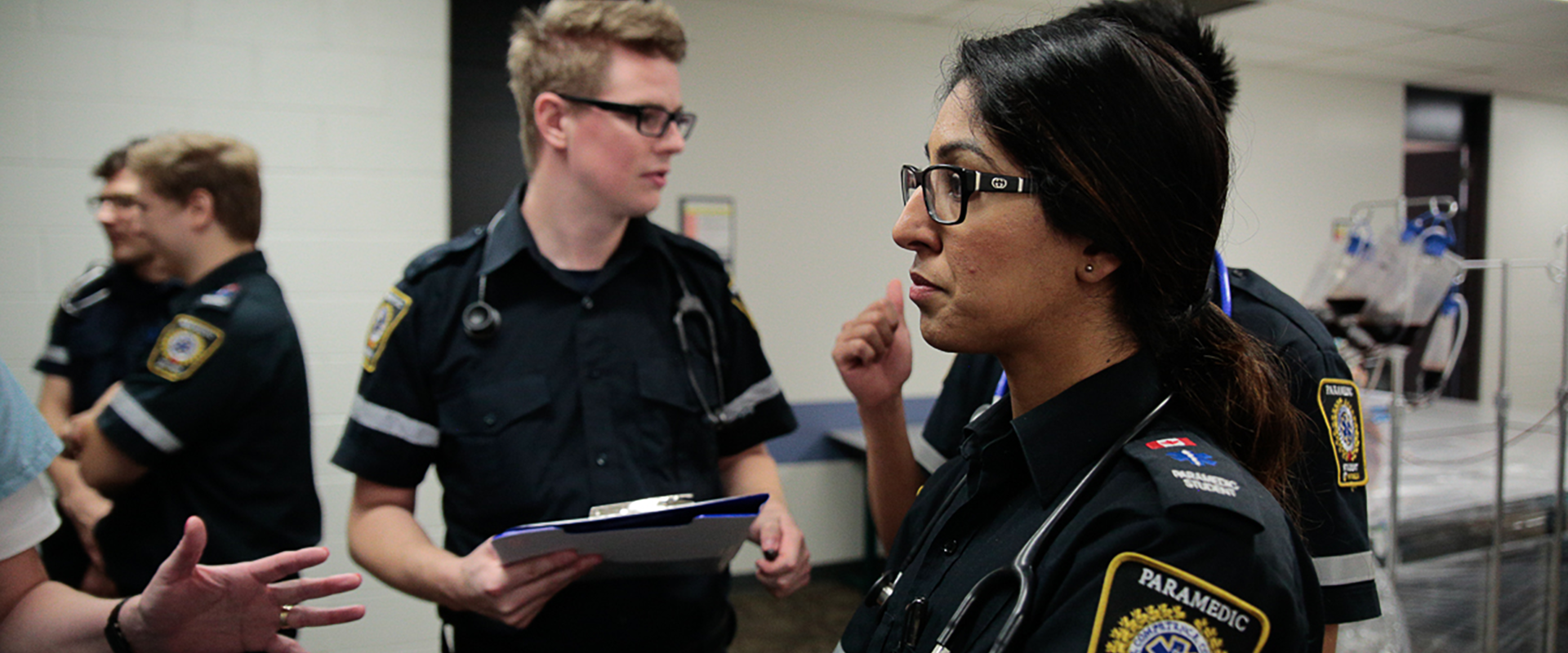 Paramedic students gathered in a room; a woman is the foreground, a man with a clipboard in the background.