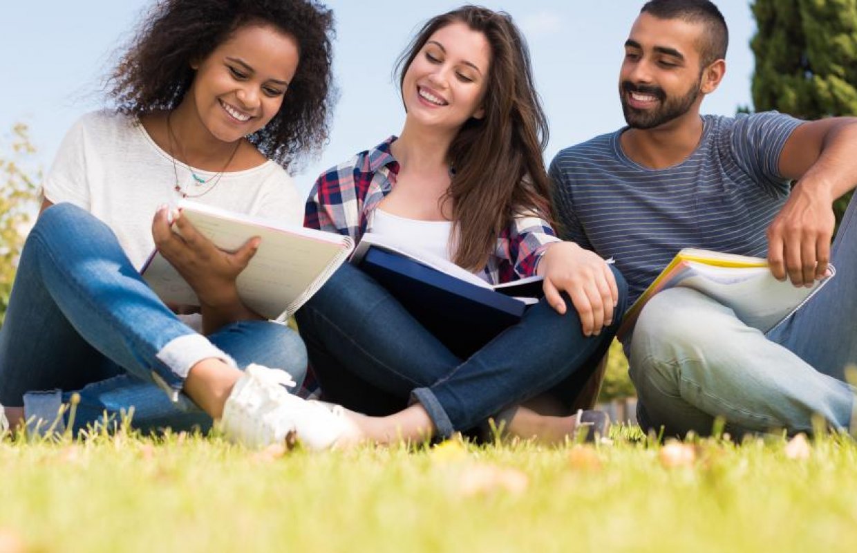 Three students sit on the grass looking at a book