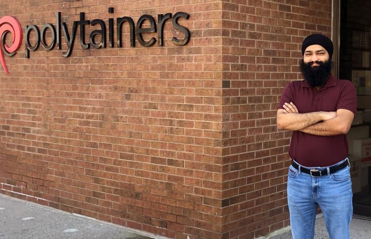 Taranjeet stands with his arms crossed beside a brick wall the the Polytainers name and logo on it.