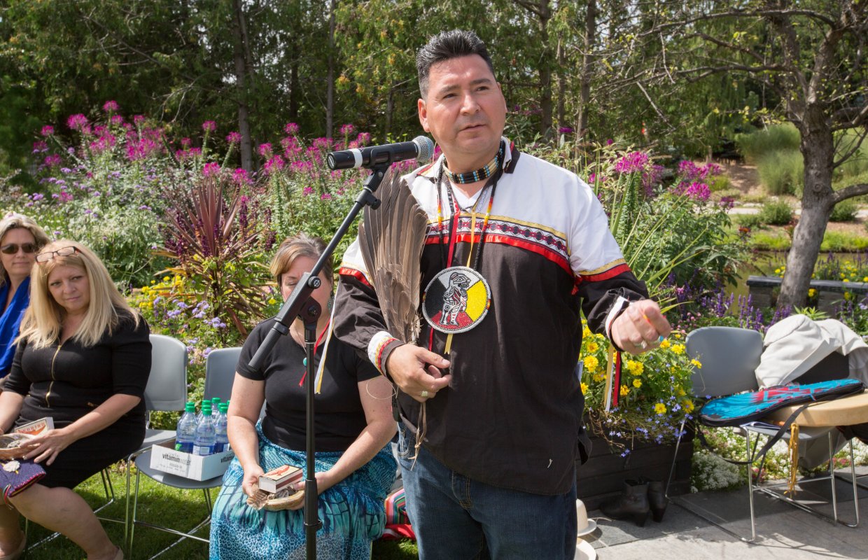 A man speaks into a microphone while addressing an audience. He wears a shirt with an Indigenous symbol on it.
