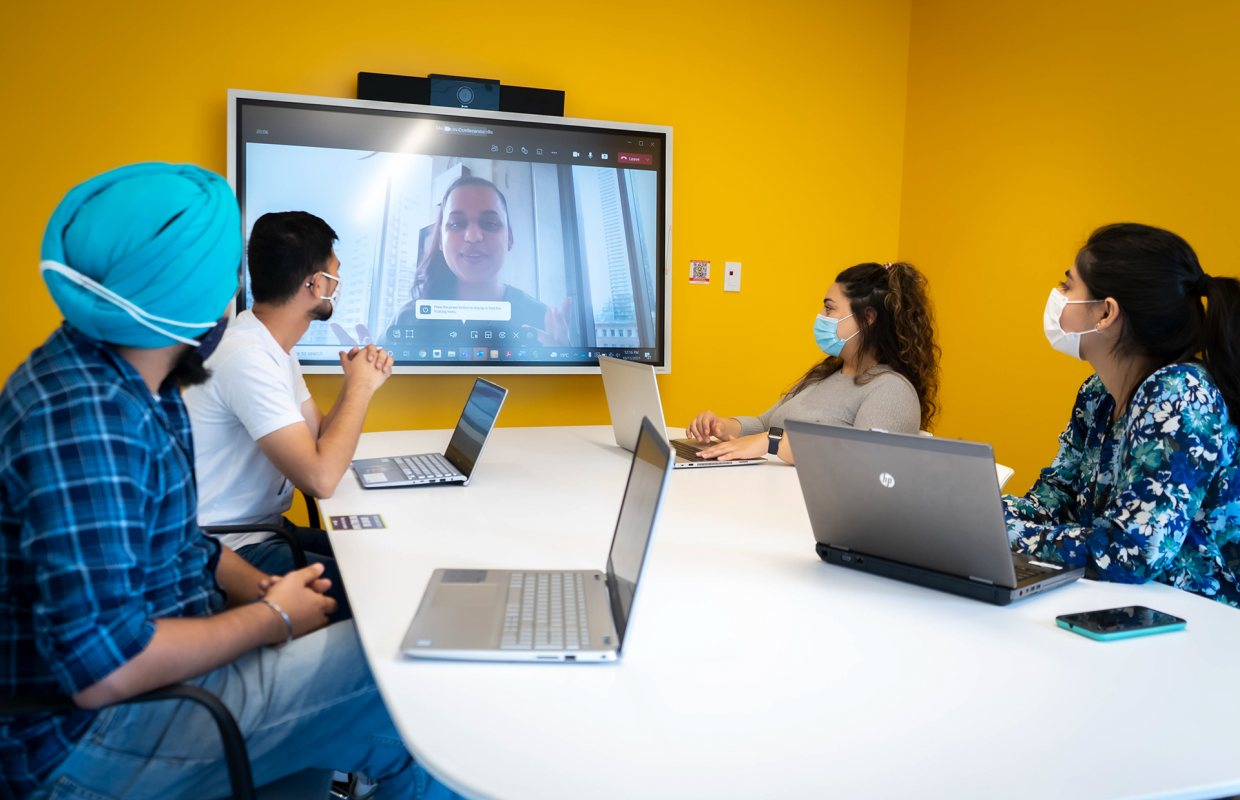 4 students sitting at a table, with a fifth student joining them virtually on a large screen. 