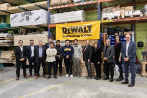 Group of people standing in front of a yellow sign that reads DEWALT Guaranteed Tough. Behind that are stacks of plywood.