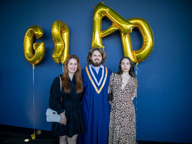 Humber graduate between two people in front of golden grad balloons