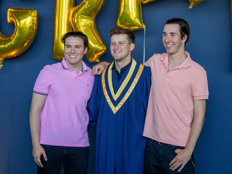 Graduate posing with two people in front of golden grad balloons