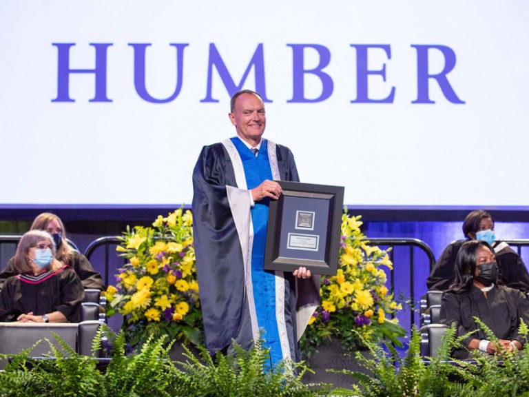 Chris Whitaker on stage holding a framed document, the display behind him says HUMBER
