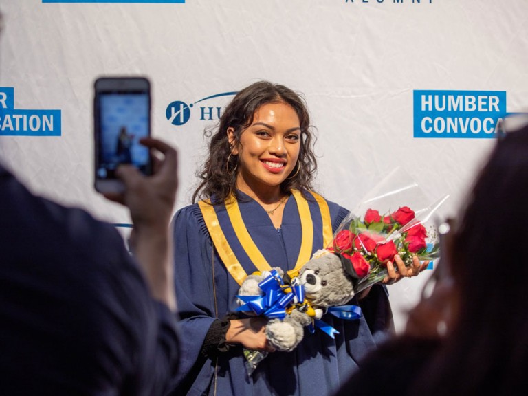 Humber graduate in front of the Humber Convocation wall holding roses