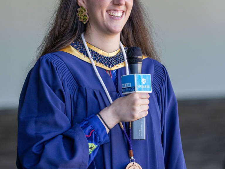 Indigenous graduate smiling with a microphone, their badge visible against their gown 