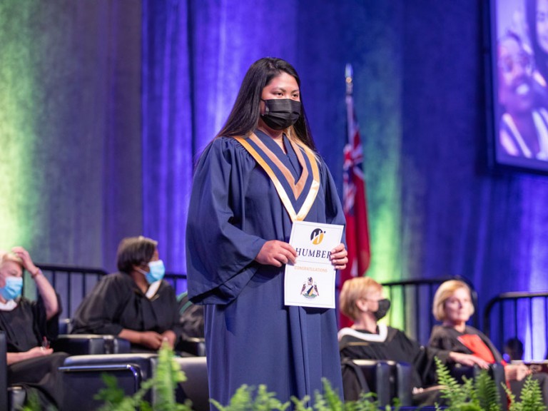 New Humber Graduate holding their diploma on stage wearing face mask