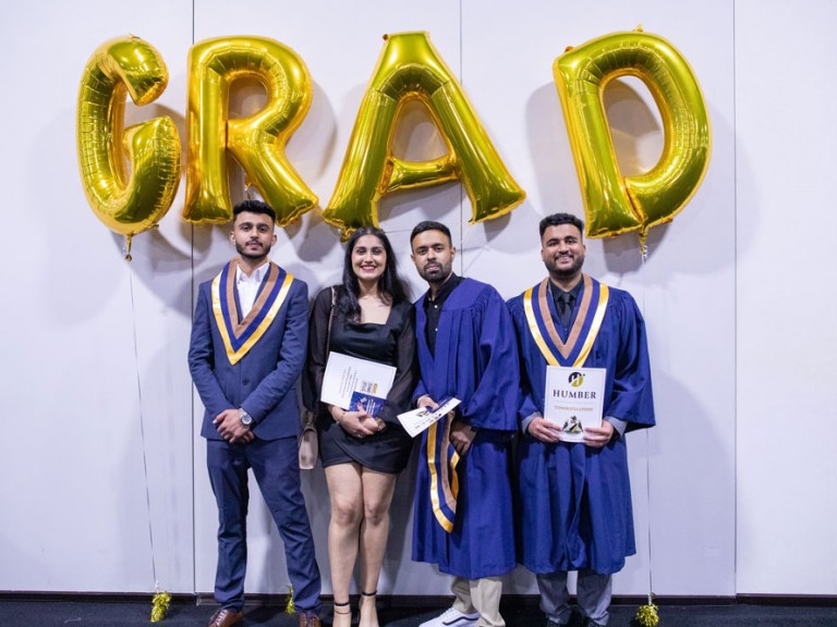 Four graduates pose in front of gold GRAD letter balloons