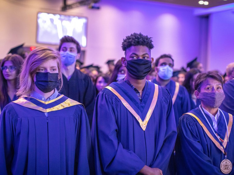 graduates in blue robes standing in front of their seats with face masks on
