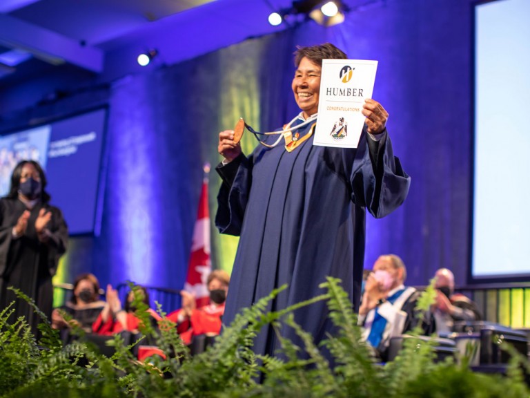 Humber Indigenous graduate smiles and holds up Indigenous badge on stage
