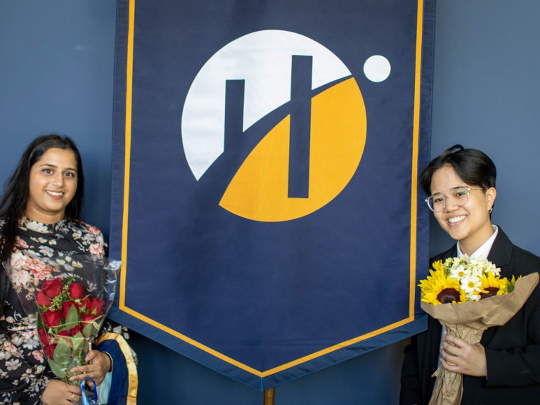 two graduates pose on each side of Humber convocation banner