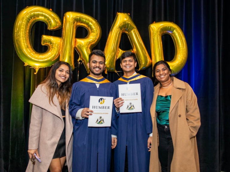 Two graduates pose with two ceremony guests in front of gold GRAD balloons