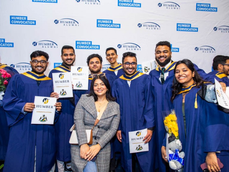 Seven graduates pose with guest in front of Humber Convocation wall