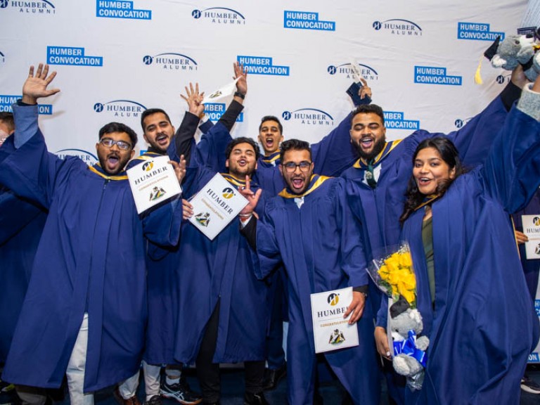 Seven graduates pose with arms up in front of Humber Convocation wall