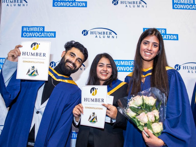 Three graduates pose in front of Humber Convocation wall