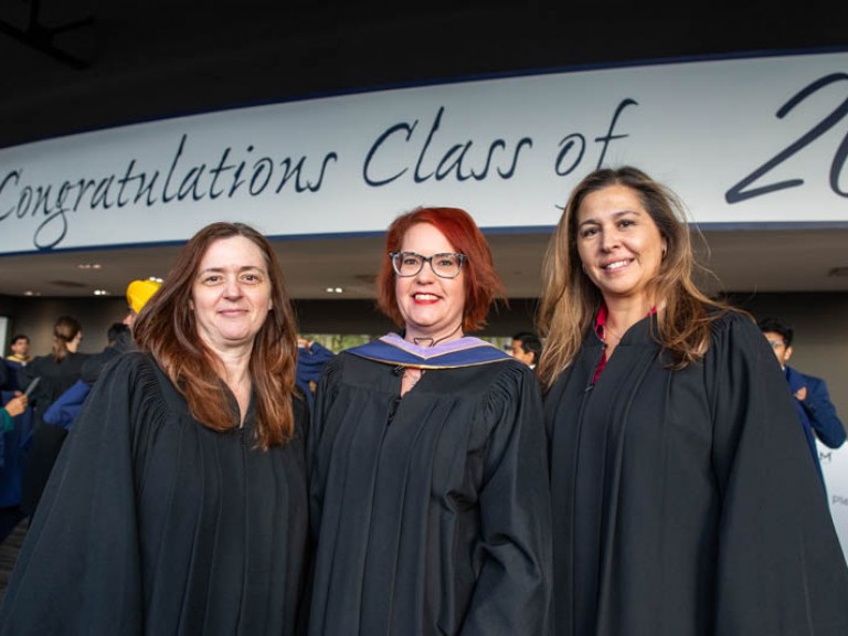 Three people in black robes pose for photo in front of Congratulations Class of 2023 sign