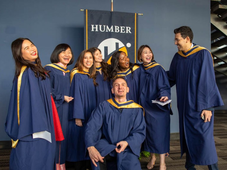 Eight graduates pose for photo in front of Humber flag