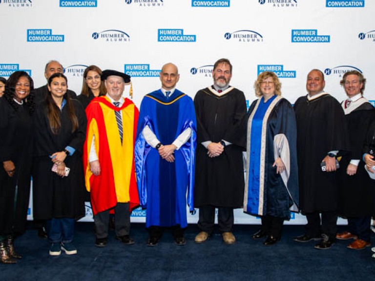 Faculty members in robes talk picture in front of Humber Convocation wall