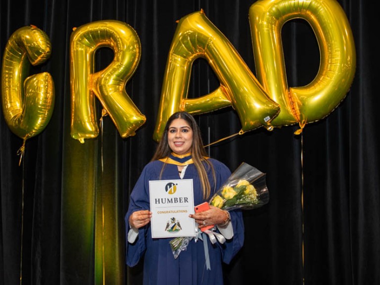 Graduate holds flowers and certificate in front of gold GRAD balloons
