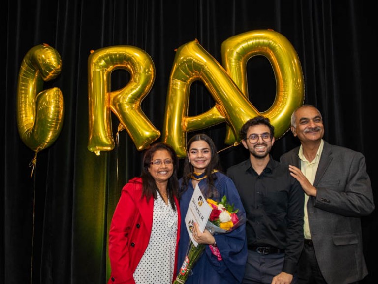 Graduate takes photo with three family members in front of GRAD balloons