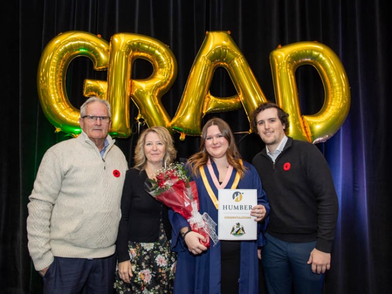 Graduate poses for photo with three family members in front of GRAD balloons