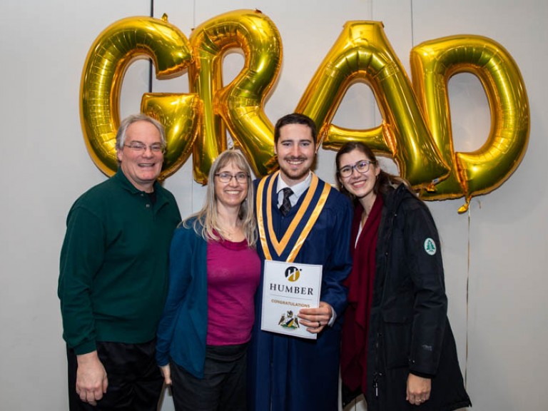 Graduate poses for photo with three family members in front of GRAD balloons