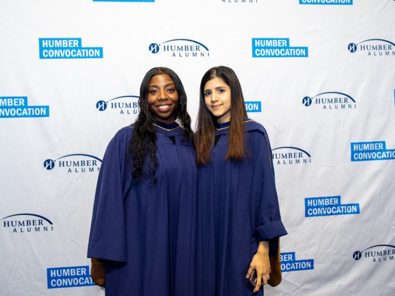 Two graduates pose for photo in front of Humber convocation wall