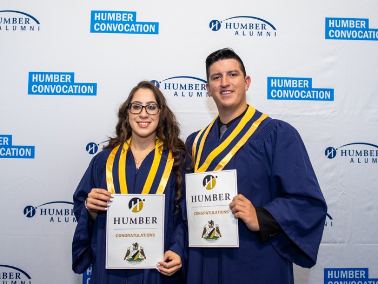 Two graduates take photo in front of Humber convocation wall