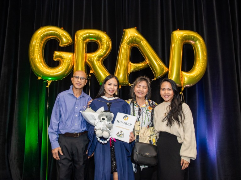 Graduate takes photo with three family members in front of GRAD balloons