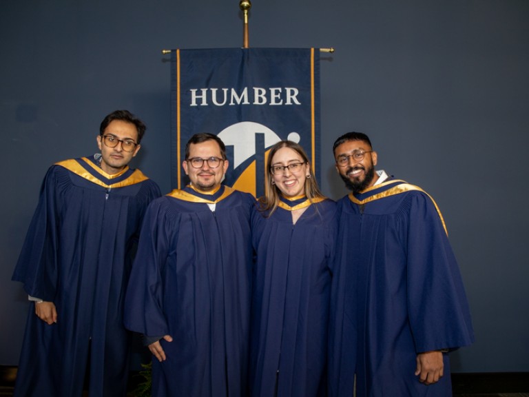 Four graduates take photo in front of Humber flag