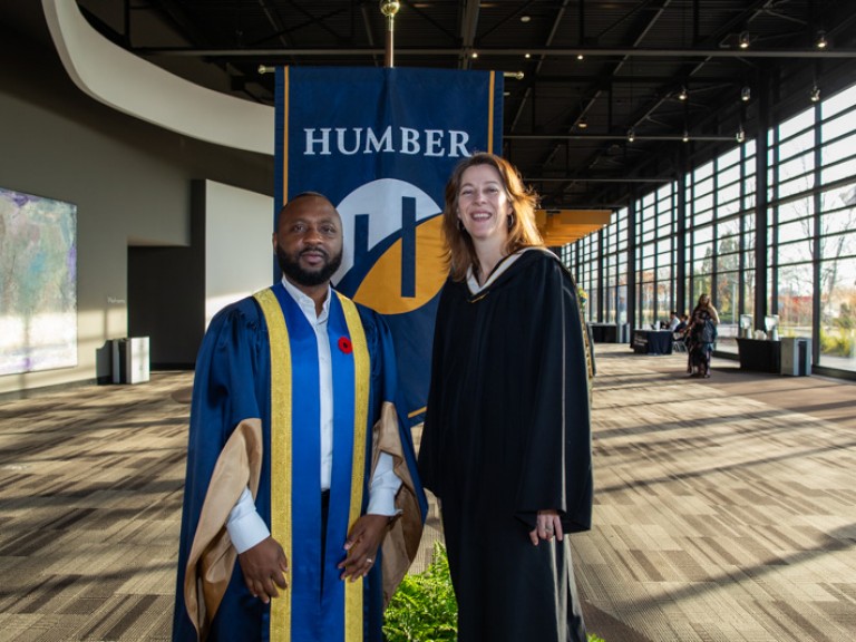 Two people in ceremonial robes take photo in front of Humber flag