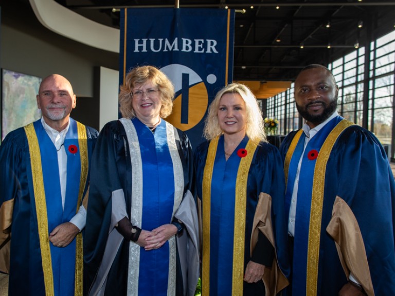 Four people wearing ceremonial robes including Humber president take photo in front of Humber flag