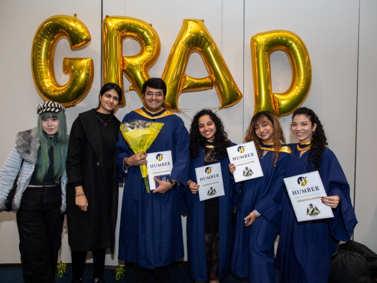 Four graduates and two guests take photo in front of GRAD balloons