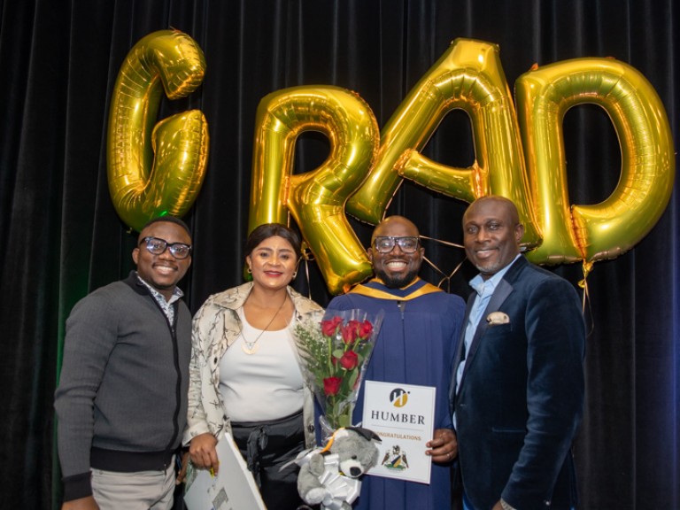 Graduate takes photo with three family members in front of GRAD balloons