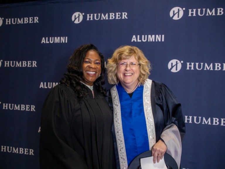 Humber president Ann Marie Vaughan takes photo with person in black robe in front of Humber Alumni wall