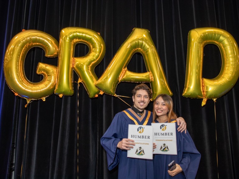 Two graduates take photo in front of gold GRAD balloons