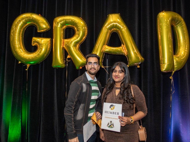 Graduate takes photo with guest in front of gold GRAD balloons