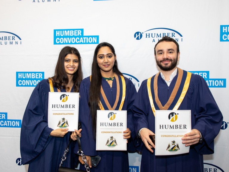 Three graduates take photo in front of Humber convocation wall