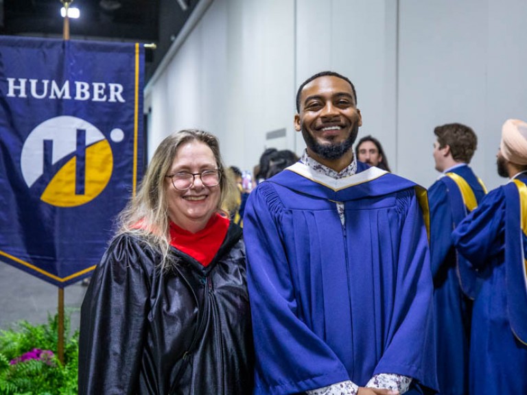 Graduate posing with another person in front of Humber flag