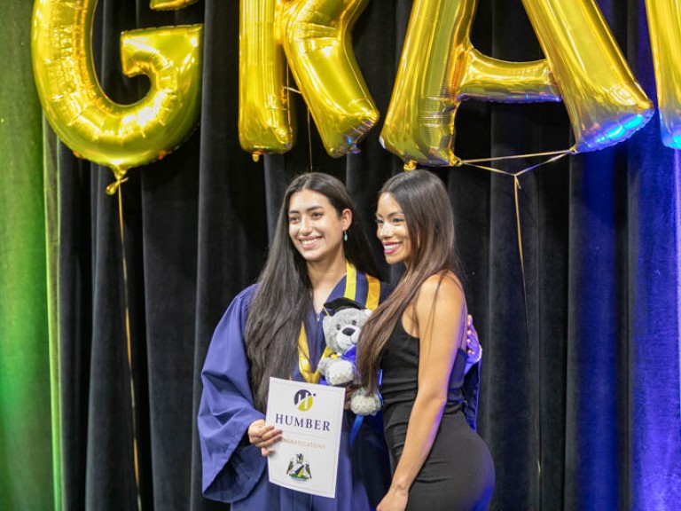 Graduate posing for photo with ceremony guest in front of gold GRAD balloons