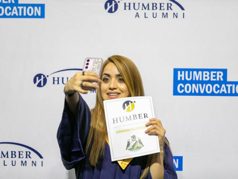 Graduate taking a selfie with her congratulations certificate in front of Humber convocation wall