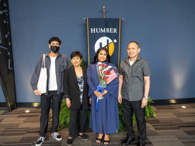 Graduate holding roses posing for photo in front of Humber flag with family