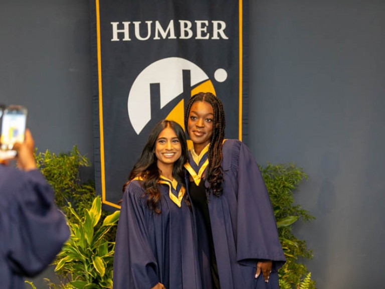 Two graduates posing for photo in front of Humber flag