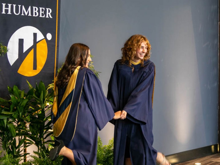 Two graduates holding hands for photo in front of Humber flag