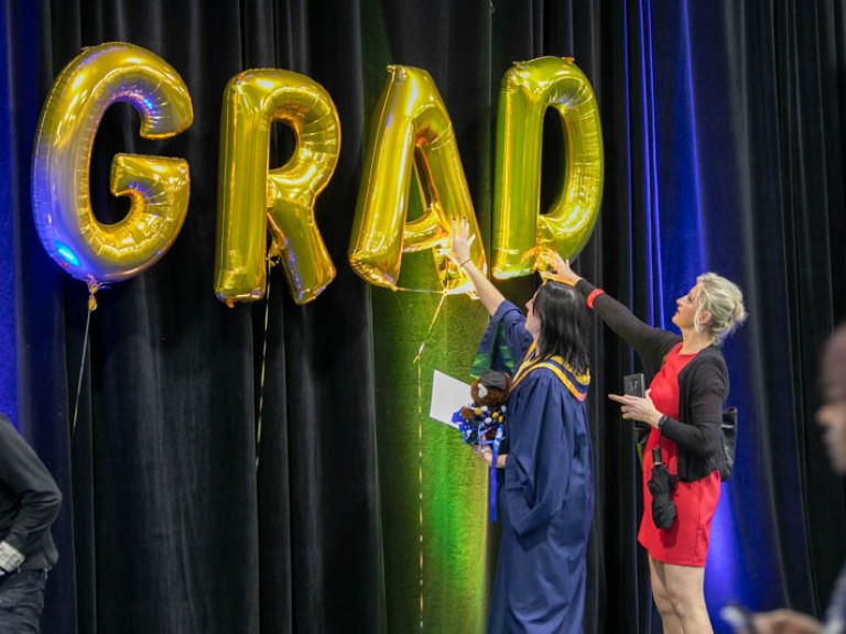 Graduate and ceremony guest touching gold GRAD balloons